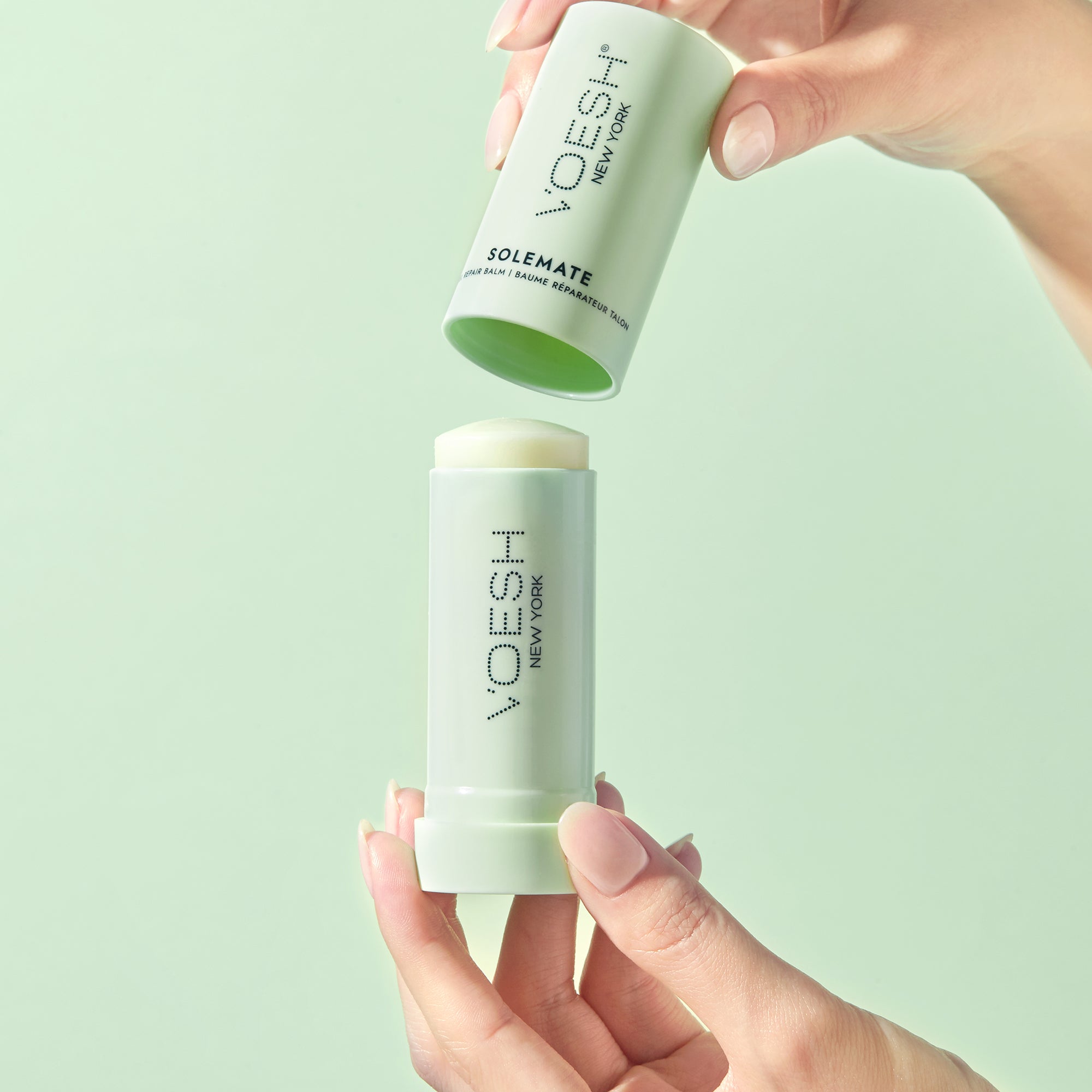 Model’s hands taking the cap off of Solemate Heel Repair Balm on a green background.